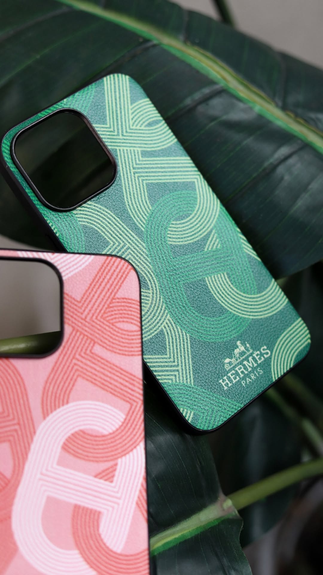 Two phone cases, one green with a pattern and the other pink with a logo, on a dark green surface.